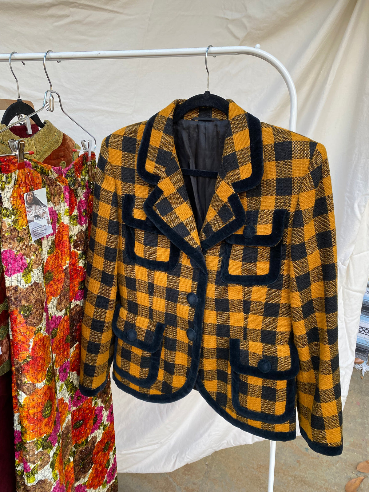 A vintage yellow and black buffalo plaid wool coat with black trim, displayed on a white clothing rack over a colorful floral quilt.