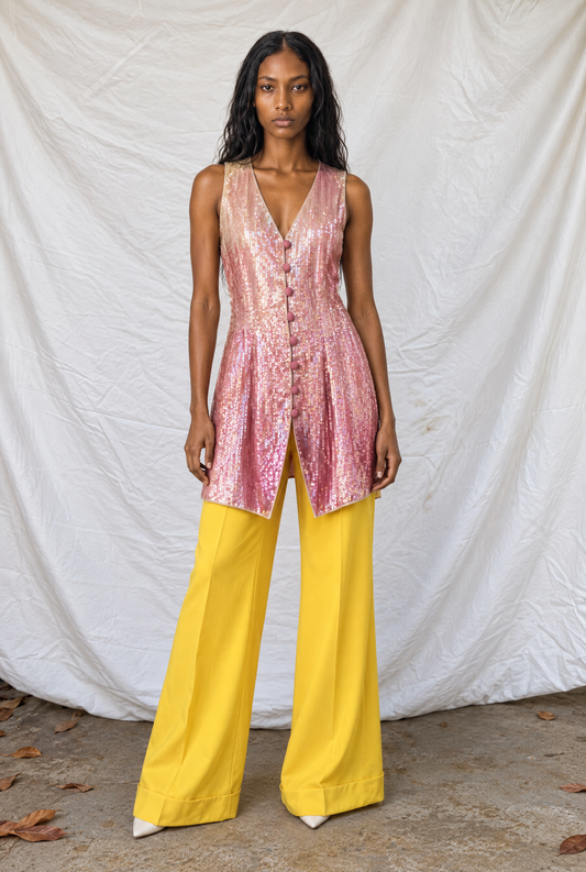 Model wearing a pink ombre sequin vest with wide-leg yellow trousers against a white drape