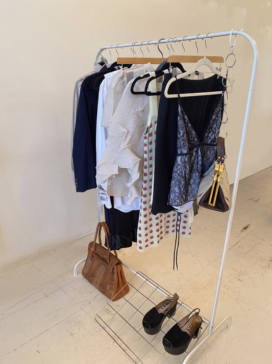 A white clothing rack holding a navy blazer, ivory pleated top, apple-print dress and lace camisole, with a croc-embossed handbag and black platform slingbacks below.