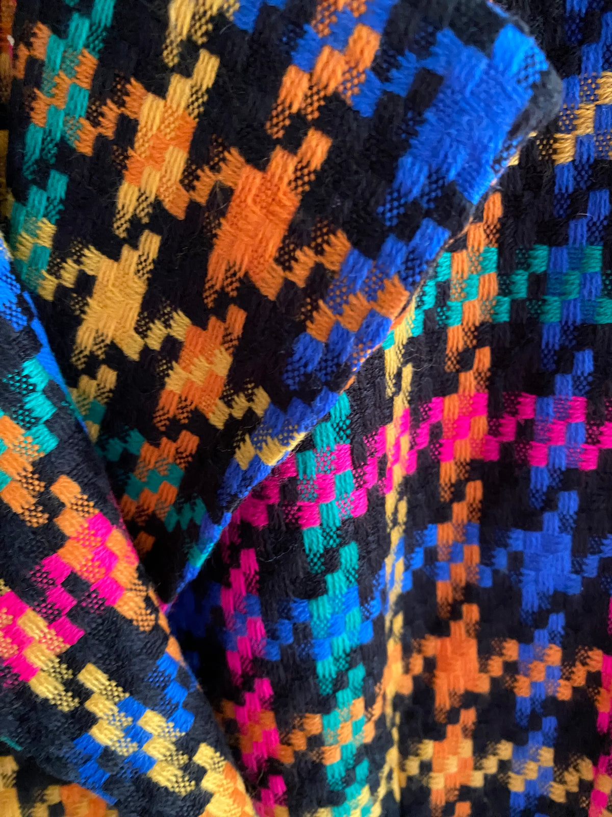 A close-up of a colorful woven textile with a geometric pixelated pattern in bright blue, orange, pink, teal, and yellow over a black base.