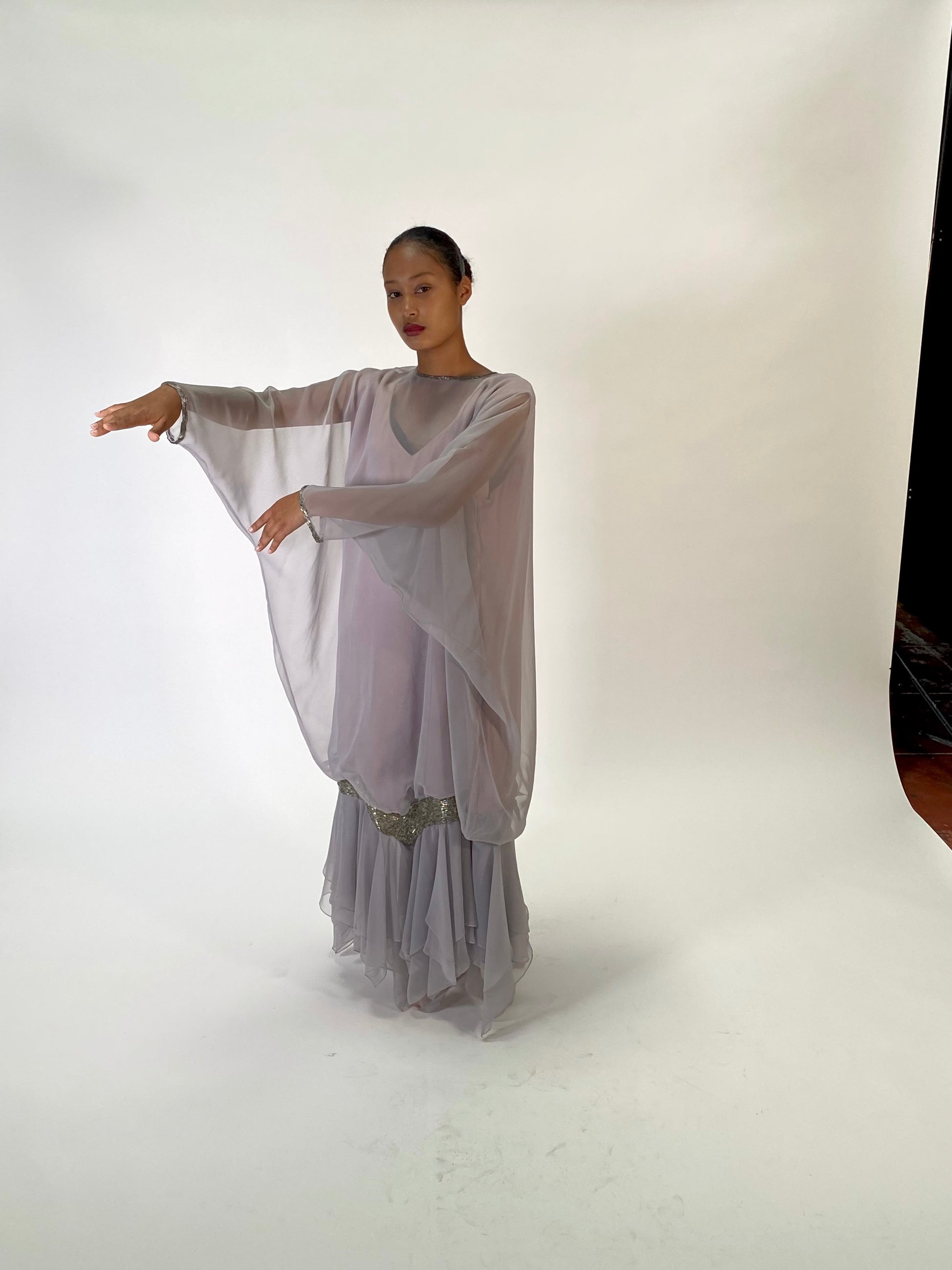 A model wearing a flowing, semi-sheer light lavender-gray dress with wide draped sleeves and a pleated skirt, posing in a studio setting against a white backdrop.