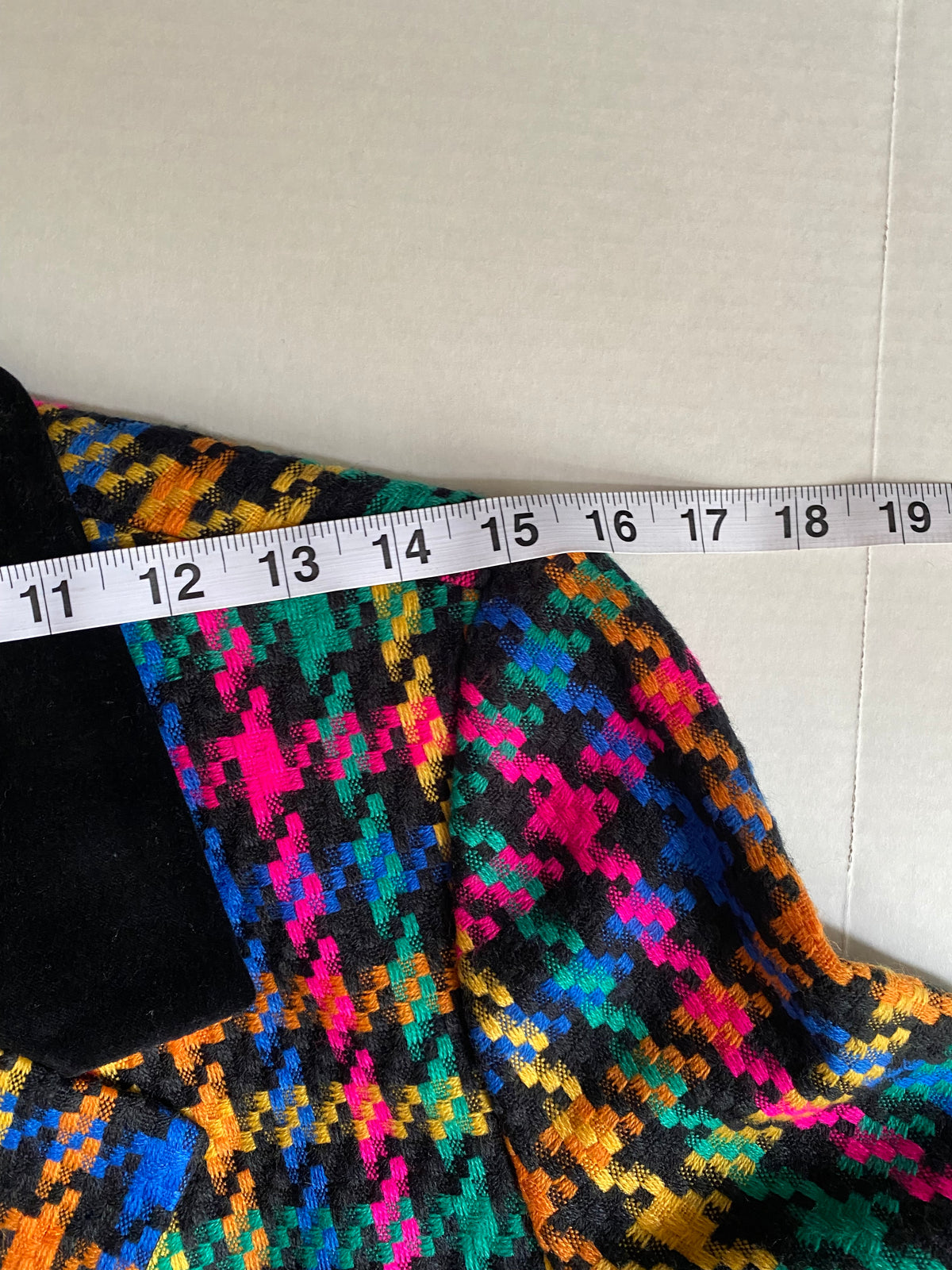 A close-up of a colorful patterned fabric garment with a black velvet-like collar, shown next to a white measuring tape to indicate size. The fabric features a vibrant, multi-colored geometric houndstooth-like pattern in pink, orange, yellow, green, and blue on a black background.