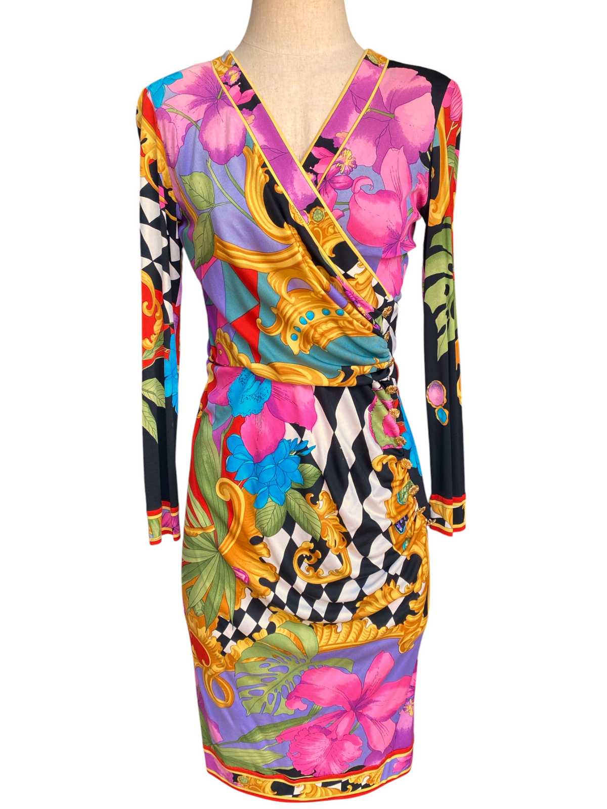 A long-sleeve wrap dress displayed on a mannequin torso, featuring a vibrant multicolor print with pink flowers, gold baroque motifs, black-and-white checkers, and green leaves.