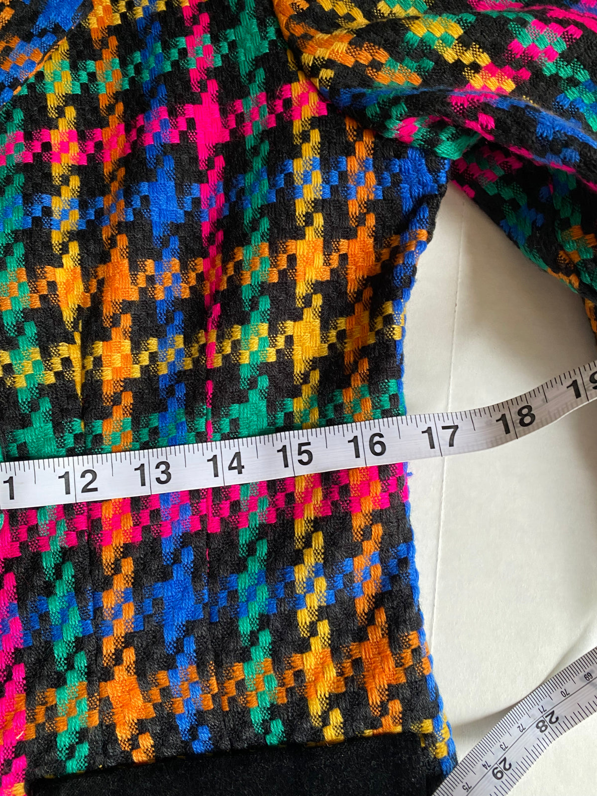 Close-up of a colorful knit garment with a geometric multicolor pattern on a black base, shown with a measuring tape across the waist area on a white surface.