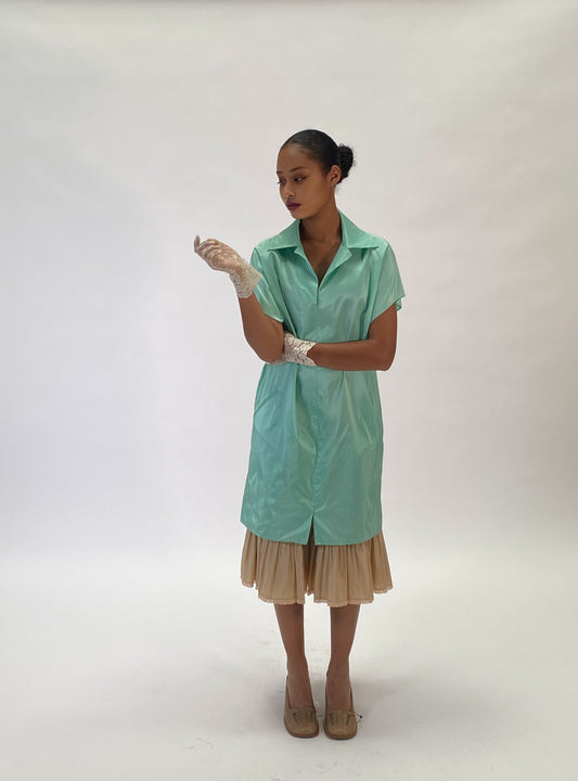 A model stands against a white studio backdrop wearing a mint green short-sleeve collared shirt dress with a beige pleated skirt, white gloves, and beige heels.