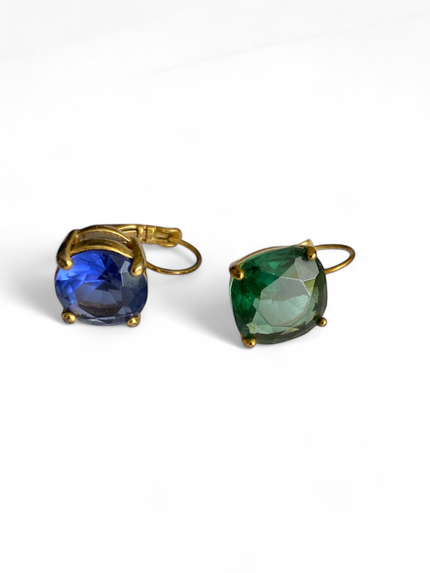 Pair of vintage-inspired earrings with mismatched faceted glass stones—one in sapphire blue, one in emerald green—set in gold-tone prongs on a white background.