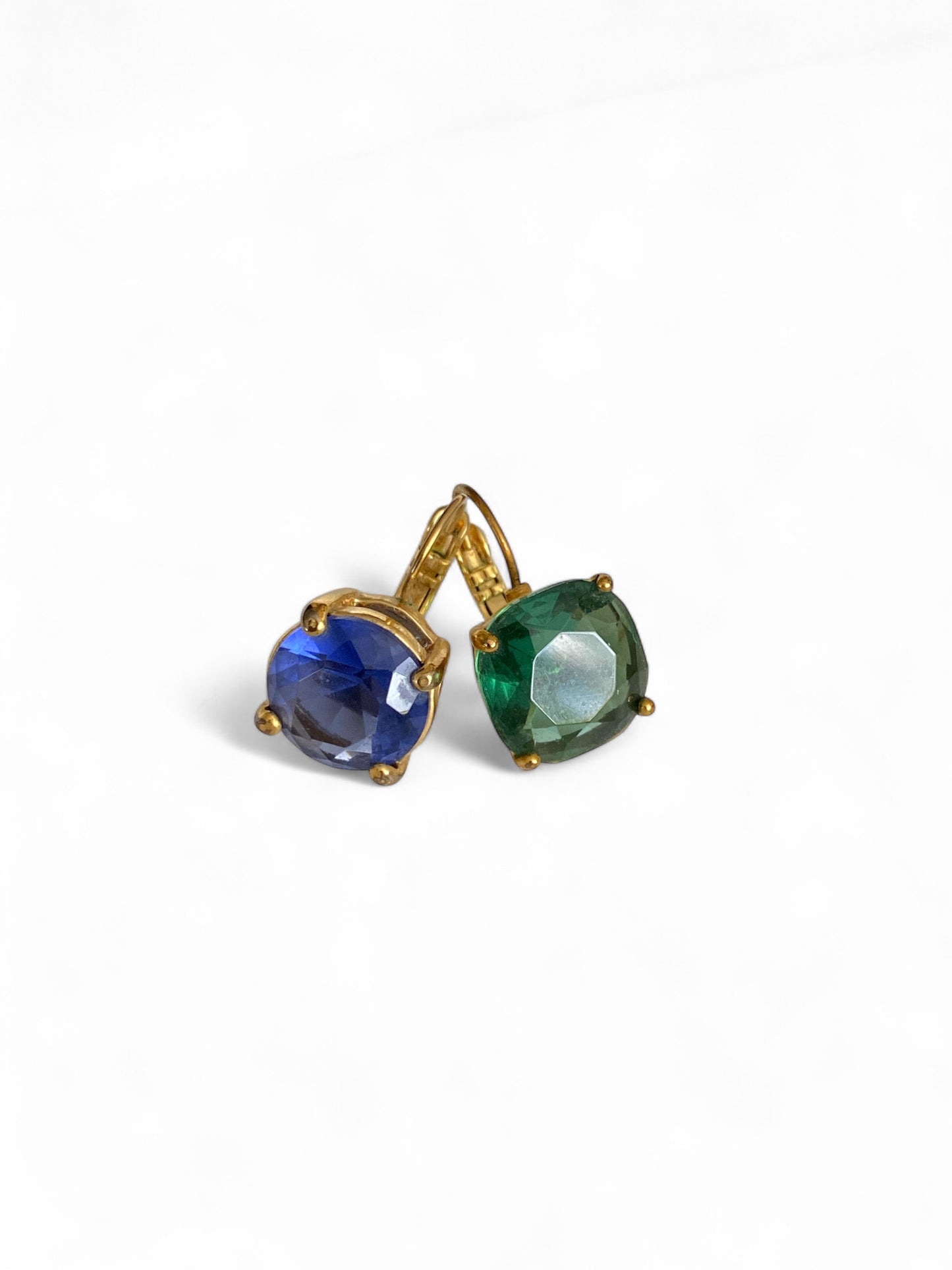 Close-up of mismatched glass stone earrings with cushion-cut emerald and sapphire colored stones, displayed in a high-end editorial style.