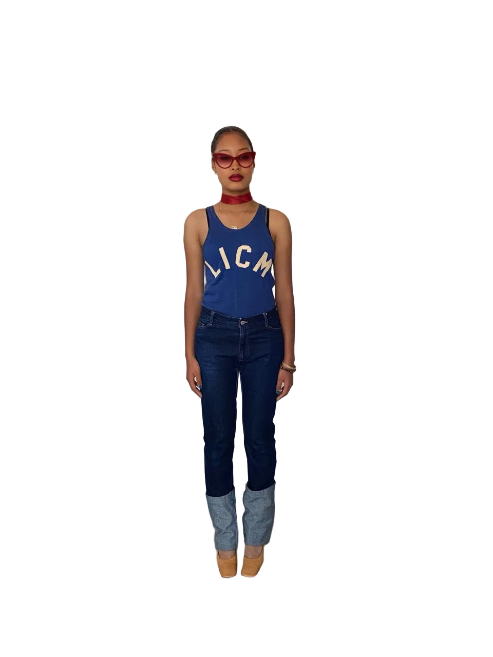 A model stands in a boutique wearing a blue tank top with 'LICMA' printed in white, dark blue jeans with light blue cuffs, and tan boots. To the left, a mannequin wears a black dress and white cowboy boots, and to the right, a clothing rack is visible against a white wall.