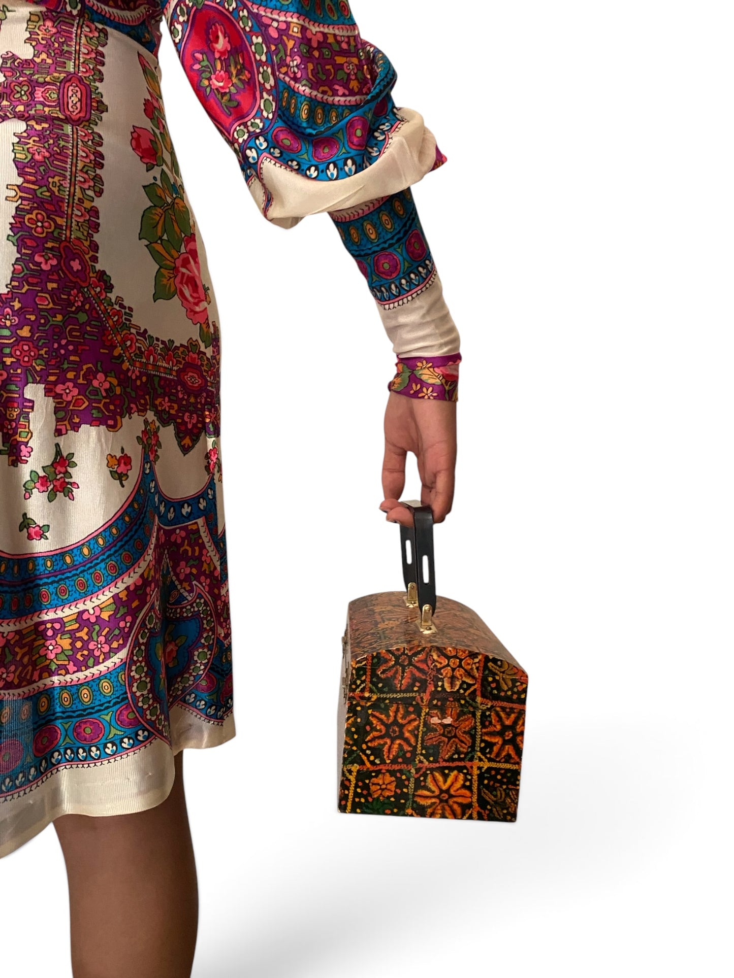A person wearing a cream-colored dress with a vibrant, multicolored floral and paisley print holds a small, matching patterned handbag with a black handle against a plain white background.