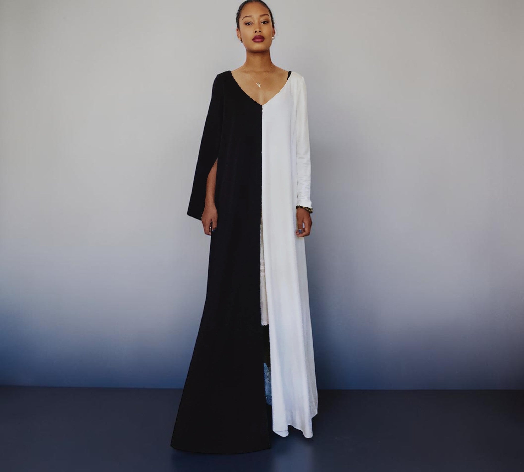 A woman modeling a long, split-color dress with one side in black and the other in white, standing against a gray studio background.