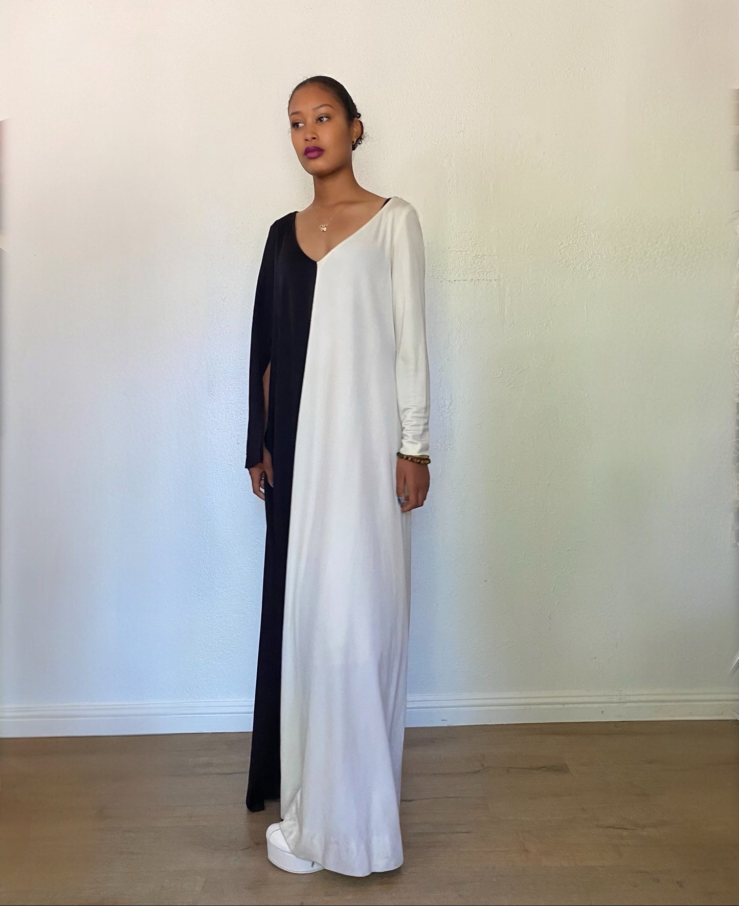 A woman modeling a long, split-color maxi dress with one side in solid white and the other in solid black, standing against a plain off-white wall.