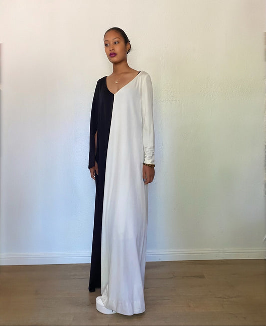 A woman modeling a long, split-color maxi dress with one side in solid white and the other in solid black, standing against a plain off-white wall.