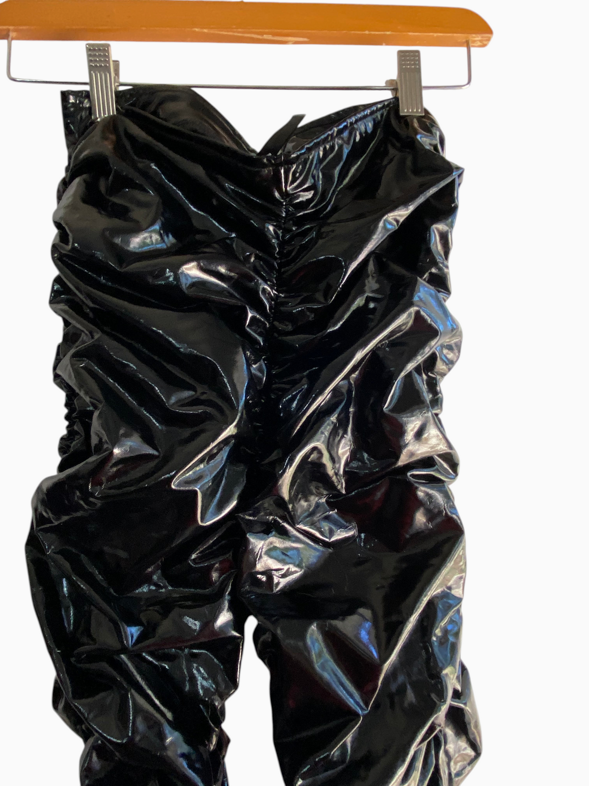 LaliSimone black shiny PU ruched pants with a form-fitting design and ankle slits hanging on a wooden hanger.